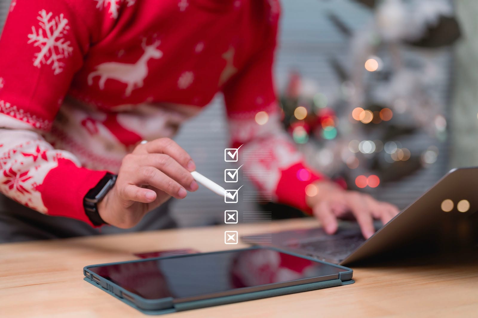 크리스마스 니트를 입은 남성이 태블릿PC와 노트북으로 체크리스트를 확인하고 있다.